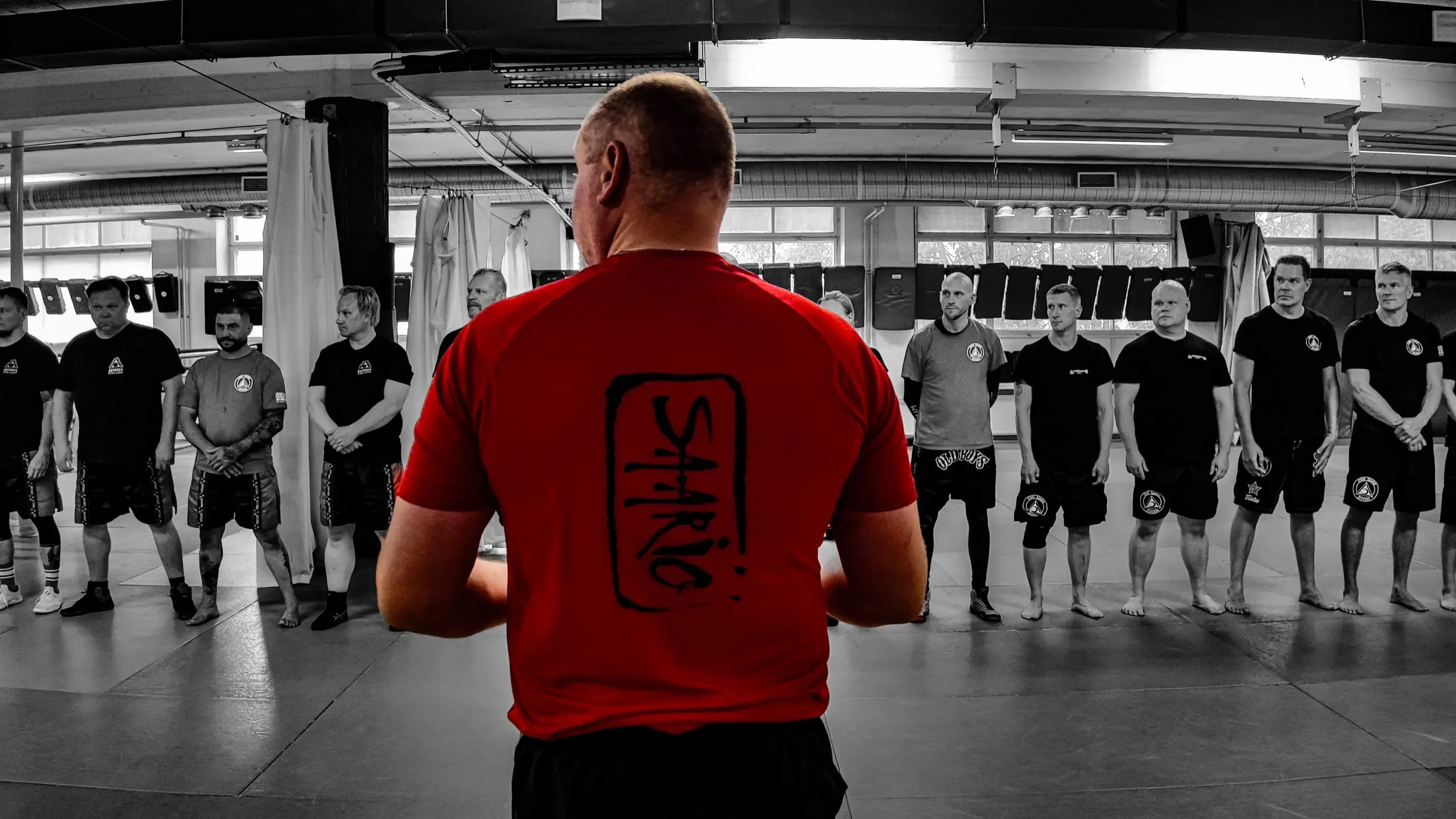 Instructor in a red Saario shirt, back to camera, addressing a disciplined line of students in the training hall