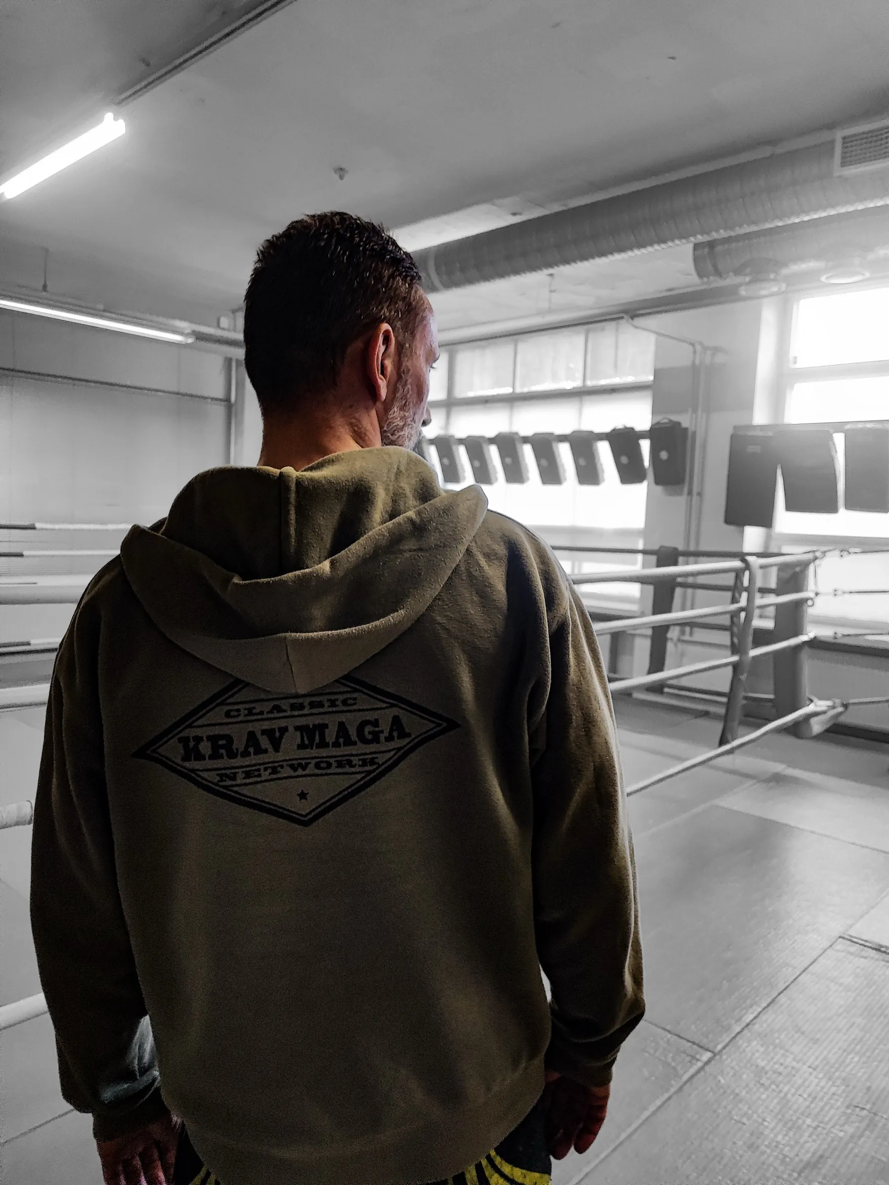 An instructor seen from behind in a Classic Krav Maga hoodie, standing at the edge of a boxing ring in a training gym